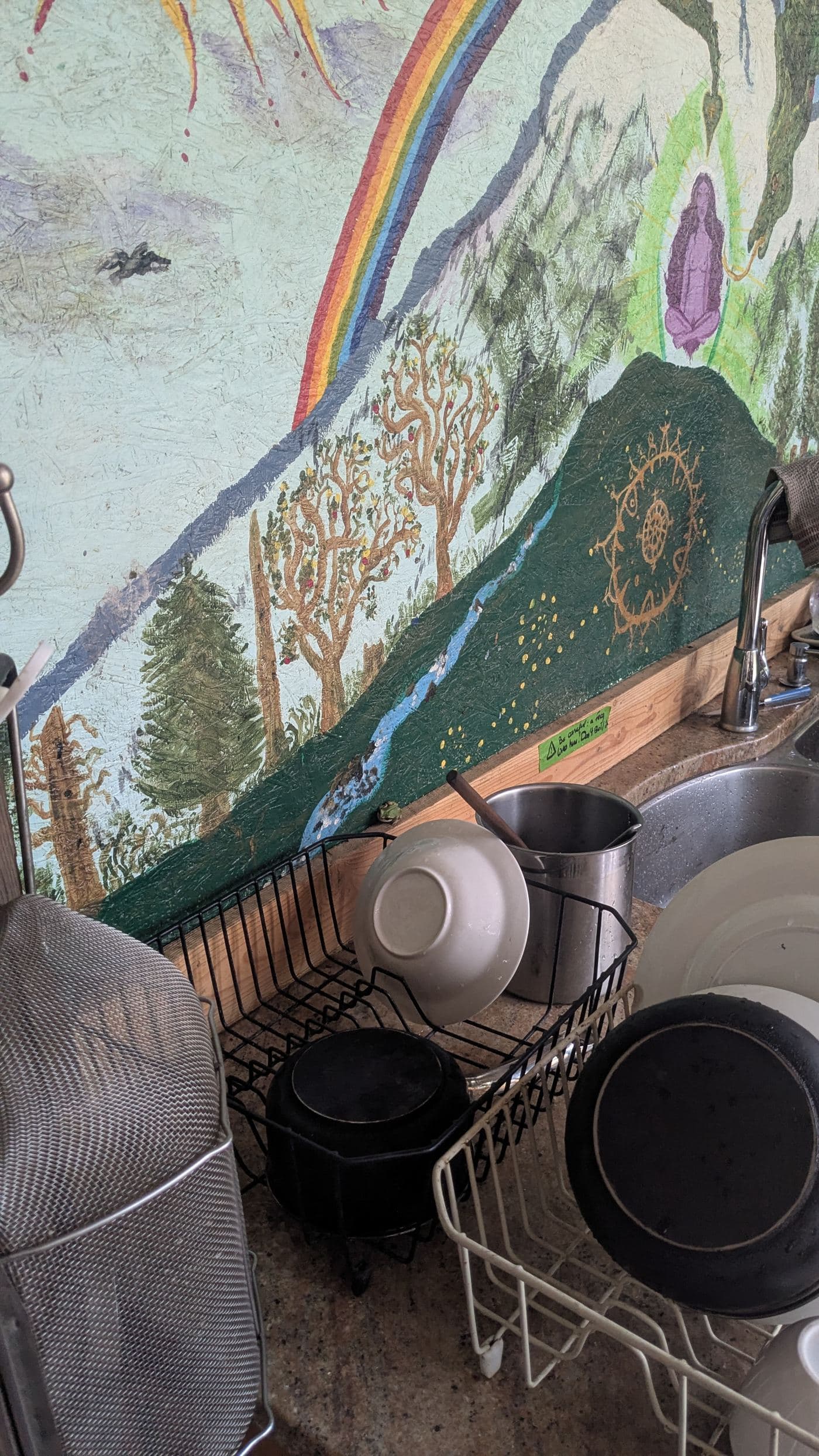 The outdoor kitchen at Entelechy, with a mural of a rainbow, orchard trees and a creek painted above the sink and drying racks